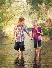 Little boy (6-7) and little girl (4-5) holding hands and walking together in small stream holding wooden stick fishing poles