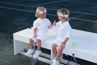 Two boys (2-3) sitting on bench with tennis rackets