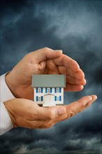 Hands holding model house in front of stormy sky.