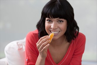 Young woman sitting on sofa and eating orange. Photo : Dan Bannister