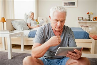 Senior man holding photo frame, woman sitting on bed in background. Photo : Rob Lewine