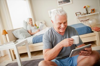 Senior man holding mug, woman sitting on bed in background. Photo: Rob Lewine