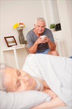 Senior man with coffee cup looking at sleeping wife. Photo: Rob Lewine