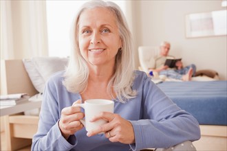Senior woman holding coffee cup with husband reading in background. Photo : Rob Lewine