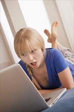 Surprised young woman lying on bed using laptop. Photo : Rob Lewine