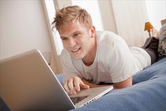 Smiling man lying on bed using laptop. Photo : Rob Lewine