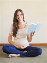 Pregnant mid adult woman sitting with crossed legs and holding book. Photo : Jessica Peterson