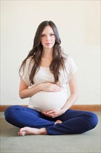 Portrait of pregnant mid adult woman sitting with crossed legs touching her abdomen. Photo :