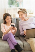 Granddaughter (8-9) and grandmother sitting on sofa and using laptop and cell phone.