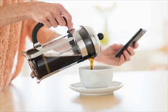 Close-up of woman pouring coffee and looking at phone.