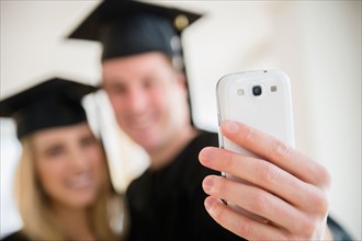 Couple of friends in graduation gowns taking photo with smartphone