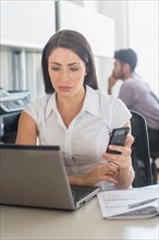 Woman working on laptop and text messaging in office, man in background.
