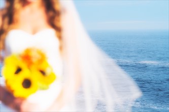 Mid section of bride holding sunflower bouquet, sea in background