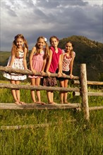 Four girlfriends (6-7, 8-9, 10-11) standing on wooden fence in grass