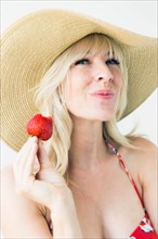 Studio portrait of blonde woman with sun hat holding strawberry