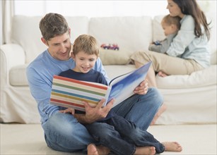 Parents with kids (12-17 months, 6-7) at home, father reading to son.