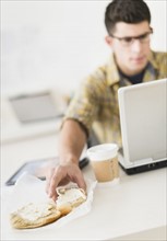 Young man eating sandwich and using laptop.