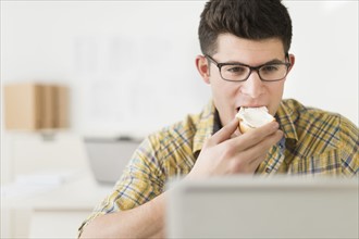 Young man eating sandwich and using laptop.