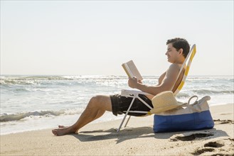Side view of man sitting on deckchair and reading book.