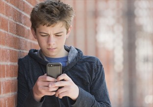 Teenage boy (16-17) leaning against brick wall texting.