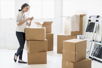 Young woman standing among boxes in new home and using mobile phone.