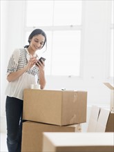 Young woman standing among boxes in new home and using mobile phone.