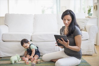 Mother using tablet pc while her son (6-11 months) playing with building blocks.