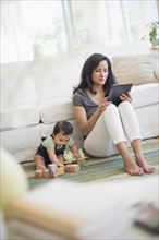 Mother using tablet pc while her son (6-11 months) playing with building blocks.