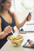 Woman eating salad and using mobile phone in office.
