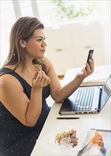 Woman eating croissant and using mobile phone in office.