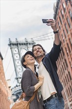 Couple taking selfie on street, Brooklyn Bridge in background. Brooklyn, New York.
