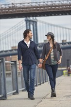 Couple walking on promenade, Brooklyn Bridge in background. Brooklyn, New York.
