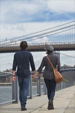Rear view of couple walking on promenade, Brooklyn Bridge in background. Brooklyn, New York.
