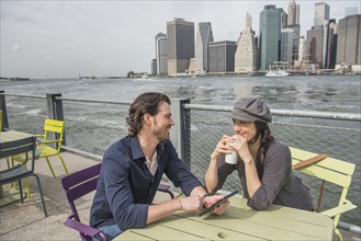Happy couple sitting and using tablet pc and drinking coffee with cityscape in background. Brooklyn, New York.