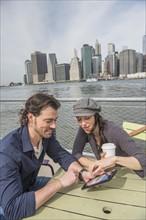 Happy couple sitting and using tablet pc with cityscape in background. Brooklyn, New York.