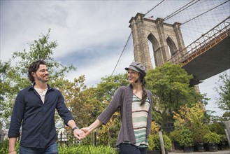 Happy couple holding hands and walking with Brooklyn Bridge in background. Brooklyn, New York.