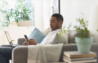 Man sitting in living room, using laptop and cell phone.
