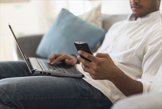 Man sitting in living room, using laptop and cell phone.