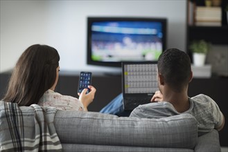 Couple sitting in living room, using laptop and cell phone.