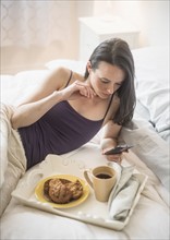 Woman having breakfast in bed, using mobile phone.