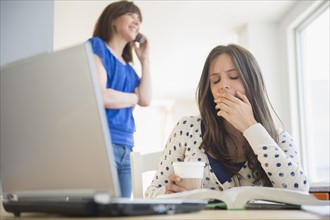 Tired teenage girl (14-15) yawning in front of textbook, mother on phone in background