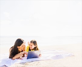 Girl (6-7) lying on beach with her mom and using tablet pc