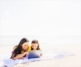 Girl (6-7) lying on beach with her mom and using tablet pc