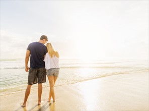 Rear view of young couple standing side by side on sandy beach, looking at sea