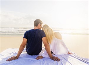 Rear view of young couple sitting on blanket on sandy beach, looking at sea