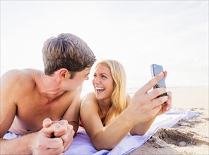 Portrait of young couple lying on blanket on beach, using smart phone