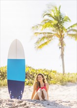 Portrait of young woman sitting near surfboard on beach