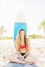 Portrait of young woman sitting on beach with surfboard in background