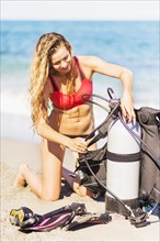 Young woman kneeling on sandy beach putting together scuba diving equipment