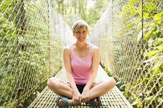 Woman sitting with legs crossed and smiling to camera on footbridge in forest